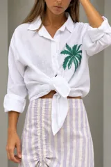 Camisa blanca de lino, con cuello clásico, cierre frontal con botones y mangas largas remangadas. Presenta un bordado artesanal de una palmera verde en el pecho.