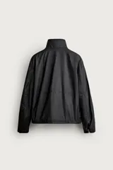 Cazadora regular fit de Zara en colaboración con Aaron Levine, confeccionada en tejido de algodón con acabado efecto encerado en color negro. Presenta cuello subido, manga larga con puño ajustable, bolsillos frontales con solapa y cierre de cremallera oculto con solapa de botones a presión.