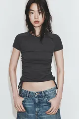 Camiseta corta de cuello redondo y manga corta, color gris oscuro, con frunces laterales ajustables mediante cordones.