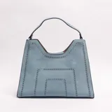Cartera tipo bolso de hombro color celeste, con diseño trapezoidal, detalle de perforaciones geométricas y un asa superior.