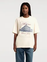 Camiseta color crema de corte amplio con estampado de paisaje costero enmarcado por columnas y la frase "Blue Horizon" en celeste.