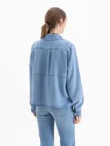Camisa de denim azul claro, manga larga, con cuello clásico y escote ajustable mediante cordón que pasa por ojalillos metálicos.