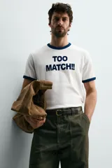 Camiseta de corte regular con cuello redondo y mangas cortas con ribetes en contraste de color. Presenta una estampa frontal con la frase 'TOO MATCH!!' en tipografía destacada.