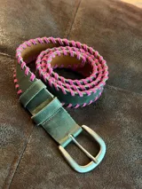 Cinturón de cuero verde con detalle de costura entrelazada en color fucsia a lo largo de los bordes. Cuenta con hebilla metálica y remaches en color plateado.