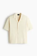 Camisa de manga corta con cuello abierto, confeccionada en tejido con textura. Presenta un corte holgado y bajo recto con aberturas laterales.
