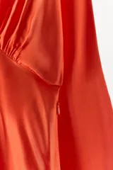Vestido largo satinado color naranja, con cuello redondo, manga larga con sisa ancha y detalle drapeado con abertura frontal en el pecho. Presenta una abertura en el bajo y cierre lateral con cremallera oculta.