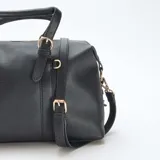 Bolso de mano estilo bowling en color negro, confeccionado en material texturizado. Presenta doble asa superior con detalles de hebillas decorativas, cierre principal con cremallera y correa larga ajustable y extraíble para llevar al hombro o cruzado. Incluye herrajes metálicos en tono dorado.