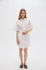 Vestido corto de algodón color blanco con diseño de broderie, cuello camisero, cierre frontal con botones, mangas cortas y lazo ajustable en la cintura.