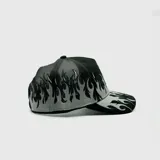 Gorra tipo trucker negra con panel frontal bordado con el logo "LA" en blanco y llamas grises. La visera también presenta bordados de llamas en blanco y gris. La parte trasera es de malla negra y cuenta con cierre ajustable.