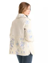 Chaqueta de corte relajado confeccionada en tejido tipo corderito color crema, combinada con paneles de matelassé en el frente y bolsillos. Presenta un delicado bordado floral en tonos azules sobre el corderito, cierre frontal metálico dorado y bolsillos de parche delanteros.