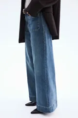 Jean de tiro alto con corte wide leg, confeccionado en denim de algodón con leve elasticidad. Presenta bolsillos de parche frontales, cierre con botón y costuras marcadas a lo largo de las piernas.