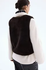 Chaleco de mujer confeccionado en tejido suave con efecto de pelo en color marrón oscuro. El diseño cuenta con cuello redondo, cierre frontal mediante botones a presión y prácticos bolsillos laterales discretos. Incluye forro interior y terminaciones con detalles revestidos.