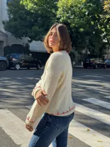 Sweater tejido color crema con cuello y puños acanalados en color naranja. Presenta un diseño bordado de un perro salchicha negro con un moño naranja.