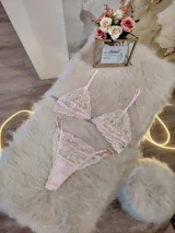 Conjunto de lencería compuesto por un bralette y una tanga, confeccionados en encaje bitono rosa y champagne con diseño floral. El bralette presenta cierre frontal con broche de gancho y breteles regulables. La tanga es de estilo colaless con laterales regulables.