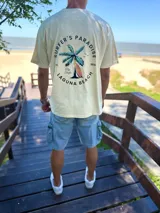 Remera oversize de color beige con estampa en la espalda de una palmera y una tabla de surf, acompañada de la inscripción "Surfer's Paradise Laguna Beach".