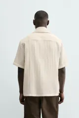 Camisa de calce relajado confeccionada en tejido con algodón de color beige. Presenta cuello bowling, manga corta, aberturas laterales en el bajo y cierre frontal con botones.