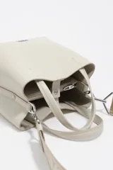 Bolso de mano de tamaño pequeño con estructura rígida, confeccionado en piel reconstituida color gris claro. Cuenta con doble asa superior, correa ajustable con logo de la marca y logo metálico en la parte frontal.