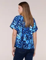Blusa de poliéster azul con estampado botánico y floral, de silueta holgada y fluido diseño. Cuenta con escote redondo elastizado con frunces y mangas cortas amplias.