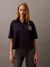Chomba azul marino de punto de algodón con cuello clásico, tapeta con botones ocultos y mangas cortas. Presenta un corte holgado y un escudo bordado con el logo de Calvin Klein en el pecho.