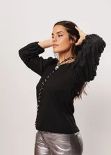 Blusa negra de tejido de punto acanalado, con cuello redondo y mangas largas abullonadas de tela semitransparente. Presenta una hilera vertical de pequeñas cuentas plateadas en el frente.