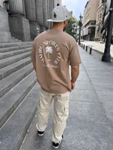 Remera color tostado con estampa en la espalda con el texto "MALIBU SPORTS CLUB" en forma circular y una palmera en el centro.