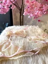 Conjunto de lencería compuesto por un bralette y una tanga, confeccionados en encaje bitono rosa y champagne con diseño floral. El bralette presenta cierre frontal con broche de gancho y breteles regulables. La tanga es de estilo colaless con laterales regulables.