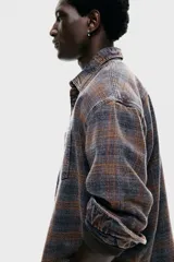 Camisa de franela de algodón suave con efecto lavado, corte relajado. Presenta cuello inglés, tapeta clásica de botones, un bolsillo superior abierto en el pecho y canesú en la espalda. Las mangas son largas con tapeta y puños abotonados, y el bajo es ligeramente redondeado. El estampado es a cuadros en tonos grises y naranjas.