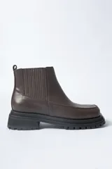 Bota corta tipo Chelsea de cuero negro, con puntera cuadrada y suela track gruesa y dentada. Presenta paneles elásticos acanalados a los lados para facilitar el calce.