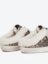 Zuecos Golden Goose modelo Super-Star Sabot, con diseño de zapatilla sin talón. Presentan exterior cubierto de glitter negro y dorado, estrella característica en el lateral, cordones y forro interior de shearling sintético.