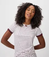 Blusa de pijama de cuello redondo y manga corta, confeccionada en tejido peach touch color blanco con estampado geométrico de corazones en tono bordó. Cuenta con detalles de encaje en los puños y un pequeño moño decorativo en el escote.
