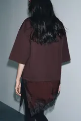 Camiseta de algodón de corte regular, color marrón oscuro o negro, con cuello redondo y manga corta. Presenta un bajo asimétrico con aplicación de encaje negro a tono.