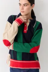 Buzo de punto con cuello polo y cierre frontal de tres botones. Presenta un diseño a rayas verticales en tonos beige y marrón, con mangas en bloques de color rojo y detalles en rosa en los puños y el ruedo.