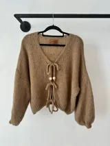 Buzo tipo saco de punto color tostado, con escote en V y cierre frontal mediante lazos con detalles de cuentas de madera. Presenta mangas largas con diseño holgado y puños ajustados.