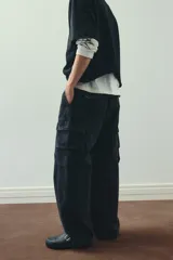 Pantalón cargo negro con efecto lavado, confeccionado en sarga de algodón suave. Presenta cintura elástica con cordón ajustable, cierre de botón a presión, bolsillos laterales, bolsillos cargo con solapa en las piernas y cordón ajustable en el bajo. Corte holgado.