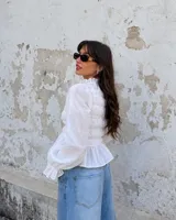 Blusa blanca con cuerpo fruncido tipo smock, cuello alto con volados y mangas largas abullonadas con puños ajustados.