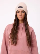 Gorro tipo beanie de punto acanalado en color beige, con parche rectangular negro con el logo de la marca en el frente.