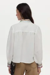Blusa de mezcla de algodón y lino color blanco, con mangas abullonadas y cierre frontal de botones. Presenta detalles de bordado geométrico a cuadros en el cuello y los puños, y un gran bordado floral en el pecho.