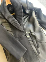 Chaqueta negra combinada de lana merino y cuero sintético, con cuello tipo chal tejido, bolsillos frontales y cinto con hebilla metálica circular.