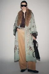 Kimono de largo midi en color verde grisáceo, con cuello de solapas amplias en pelo sintético marrón. Presenta mangas largas acampanadas y un cinturón ajustable del mismo tejido. El diseño está decorado con un delicado bordado floral en tonos tierra y rosados.