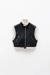 Chaleco biker negro de cuero sintético con forro de borrego beige, cierre frontal metálico, bolsillos laterales con cremallera y cinturón ajustable.