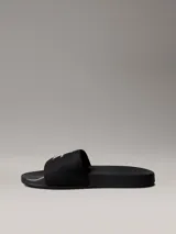 Ojotas tipo slide Calvin Klein Jeans, color negro, con banda superior de lona que presenta el logo de la marca bordado en blanco. Posee suela de EVA con plantilla ergonómica moldeada y el nombre de la marca grabado en el lateral.