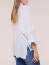 Camisa blanca de silueta holgada y textura liviana, con cuello camisero clásico, abotonadura frontal con botones en contraste y doble bolsillo de parche en el frente. Cuenta con mangas de corte dolman de largo tres cuartos con puño amplio y ruedo recto con aberturas laterales.