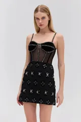 Vestido corto negro sin mangas confeccionado en terciopelo y tul transparente, con corsé estructurado con ballenas y tirantes finos. Presenta detalles de pedrería y bordados brillantes en las copas, los tirantes, las costuras frontales y a lo largo de la falda de encaje.