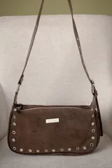 Cartera tipo baguette de gamuza color bordó, con detalles de tachas metálicas plateadas en el contorno inferior y laterales. Cuenta con cierre superior, correa ajustable y placa metálica con el logo de la marca en el frente.