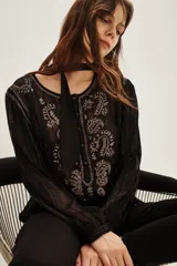 Camisa de manga larga en color negro con tejido semitransparente y detalles de hilos metalizados. Presenta un diseño con bordados florales en tonos tierra, cierre frontal con botones y un lazo decorativo en el cuello.