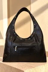 Cartera tipo hobo de gamuza color marrón chocolate, con tira de cuero decorada con tachas metálicas doradas y negras. Incluye placa metálica con el logo de la marca en la base.