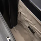 Mochila de diseño estructurado en color negro, confeccionada en material símil cuero con textura granulada. Presenta una solapa frontal con cierre metálico decorativo y logo de la marca en la parte inferior.