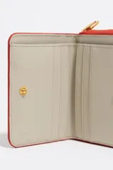Billetera pequeña de estructura rígida con acabado brillante y estampado animal print en tonos rojos y negros. Presenta cierre de cremallera perimetral, logo metálico dorado en el frente y tarjetero interior.