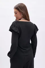 Blusa negra de manga larga con textura, cuello en V y detalle de volados en los hombros. Presenta mangas con puños elásticos y un corte recto.