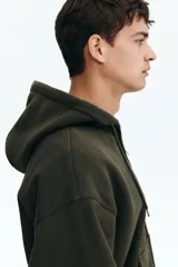 Campera con capucha y cierre frontal, color verde militar oscuro, de tejido pesado con interior cepillado suave. Presenta hombros caídos, cordón ajustable en la capucha y bolsillos canguro delanteros.